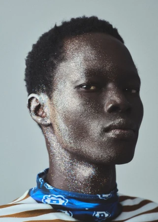 Close-up studio portrait of a dark-skinned adult male with glitter on his face and neck, wearing a striped shirt and blue bandana.