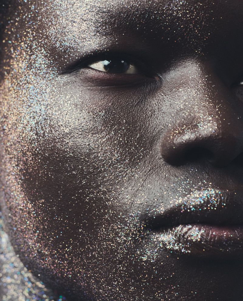 Close-up portrait of a dark-skinned person's face covered in shimmering silver glitter, highlighting their eye and lip features.