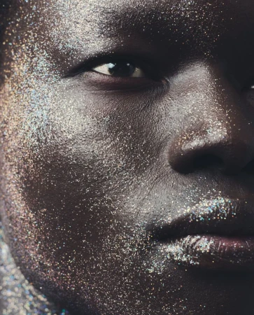 Close-up portrait of a dark-skinned person's face covered in shimmering silver glitter, highlighting their eye and lip features.