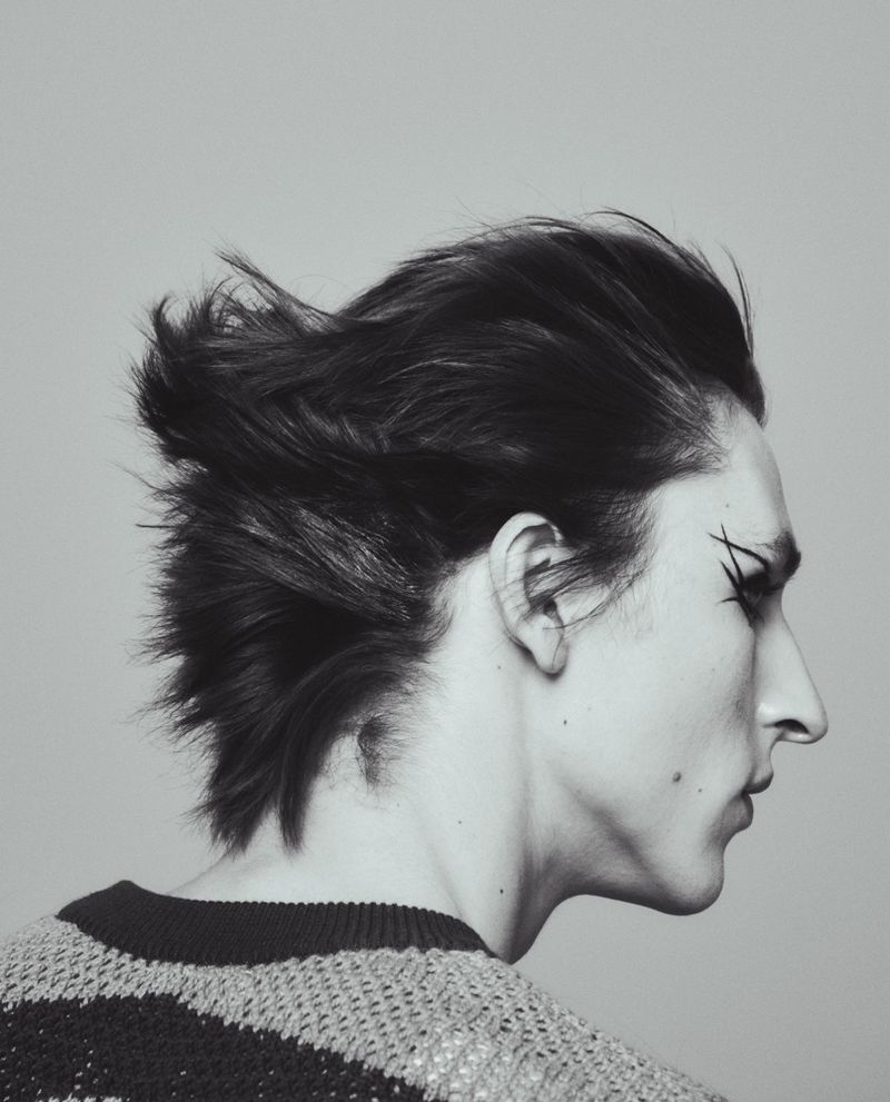 Profile portrait of a model with styled dark hair and graphic eyeliner wearing a striped sweater against a light grey background.