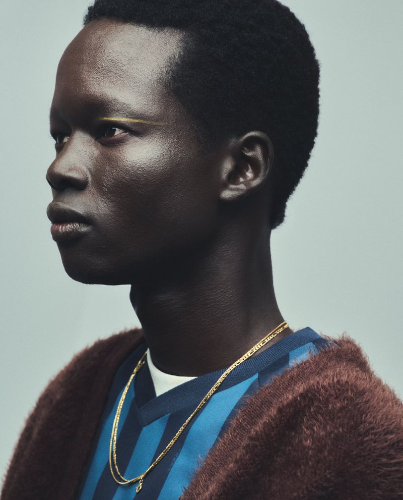 Portrait of a dark-skinned adult male model in a blue and black striped shirt and brown jacket, against a light gray backdrop.