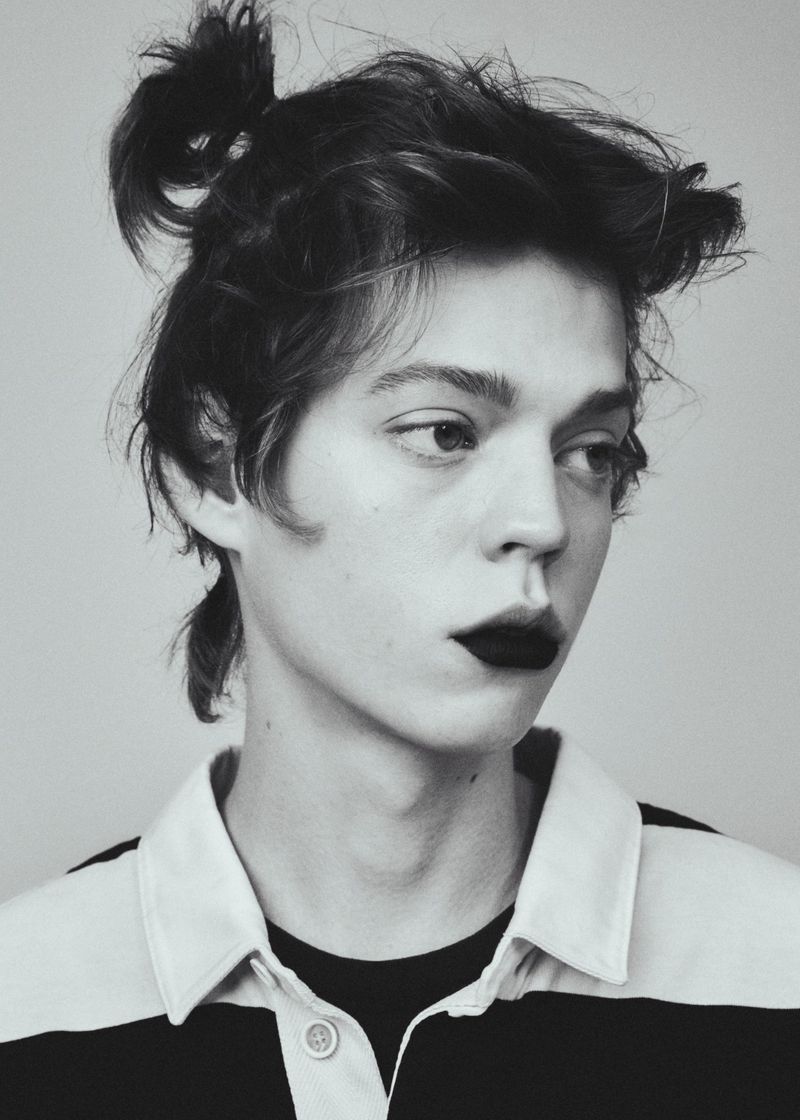 Portrait of a young adult male with hair in a bun and dark lipstick, wearing a rugby shirt against a light grey background.