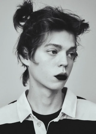 Portrait of a young adult male with hair in a bun and dark lipstick, wearing a rugby shirt against a light grey background.