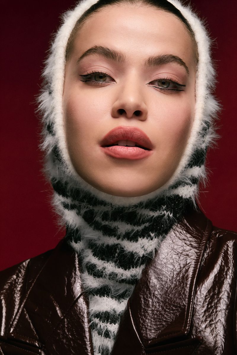 Portrait of a woman in a black and white balaclava and brown leather jacket against a red background in a studio shot.