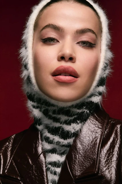 Portrait of a woman in a black and white balaclava and brown leather jacket against a red background in a studio shot.