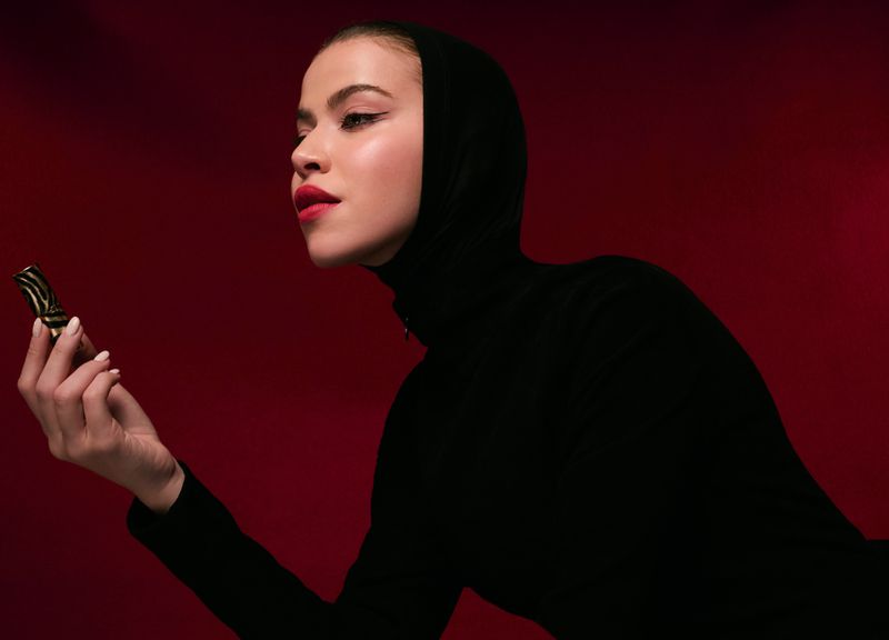 Elegant woman in black headscarf and dress holds patterned lipstick case against a red backdrop, showcasing beauty and fashion.