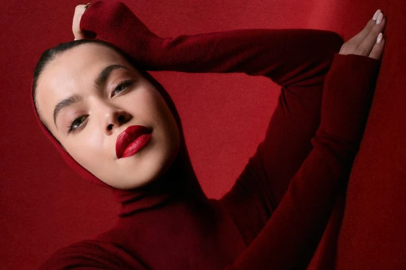 Close-up shot of a model with red lipstick and a red turtleneck sweater, posing against a matching red background in a beauty shot.
