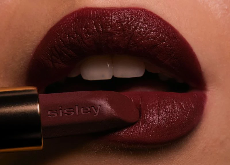 Close-up of a woman's mouth with dark red lipstick and a Sisley lipstick tube covering part of her lip in a beauty studio shot.