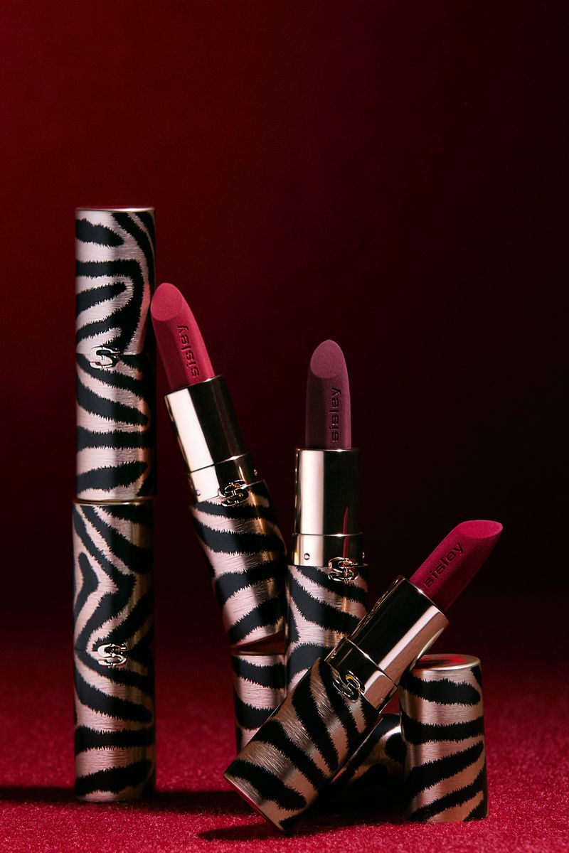 Stack of Sisley brand lipsticks with zebra print cases and red lipstick on a matching red textured surface and background.