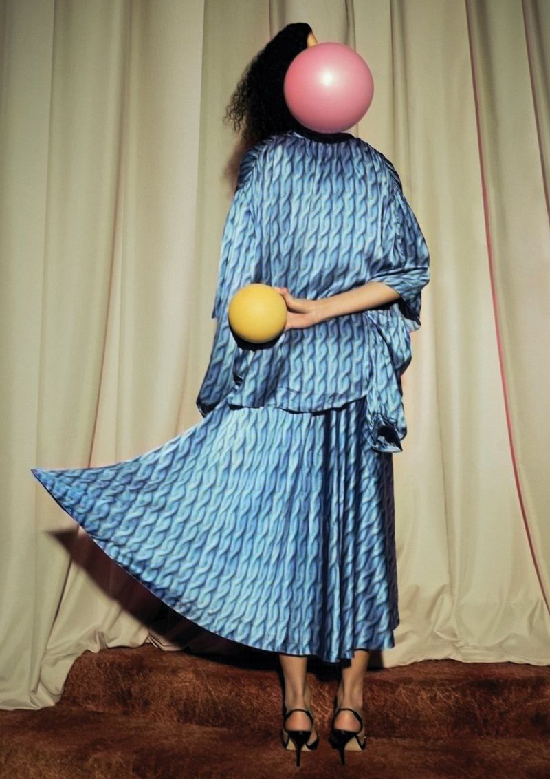 A person is wearing a blue patterned dress and holding a yellow ball, with a pink ball obscuring their face, against a neutral backdrop.