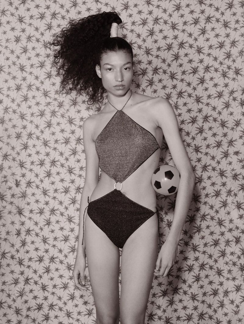 Portrait of a slender young woman in a cutout swimsuit holding a soccer ball against a patterned backdrop in a studio setting.