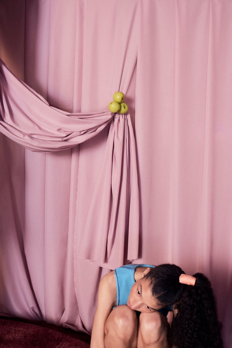 Portrait of a young woman kneeling indoors against a pink curtain backdrop adorned with green apples, showcasing a blend of fashion and art.