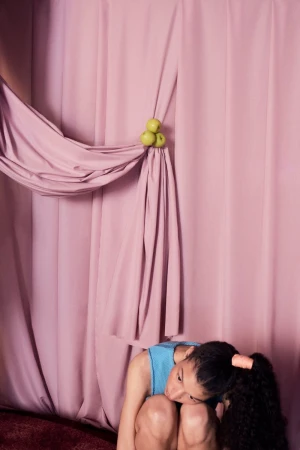 Portrait of a young woman kneeling indoors against a pink curtain backdrop adorned with green apples, showcasing a blend of fashion and art.