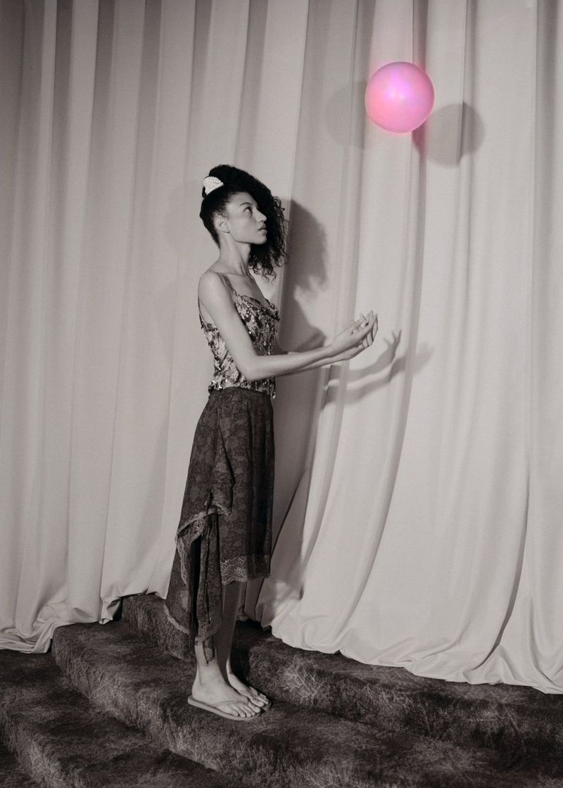 Woman in a floral top and dark skirt stands on carpeted steps reaching towards a pink balloon, with a white curtain backdrop in a studio setting.