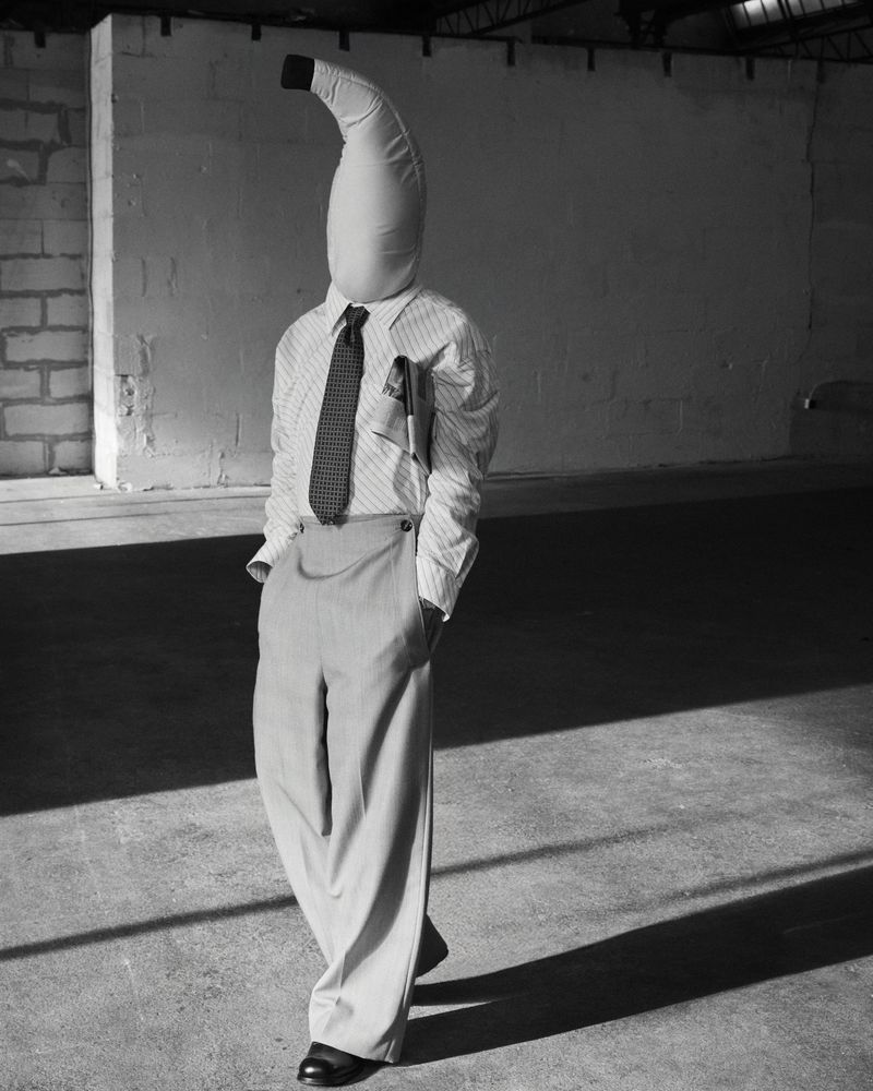 Person wearing a banana costume with a shirt, tie, and trousers stands in a concrete indoor space with a brick wall.
