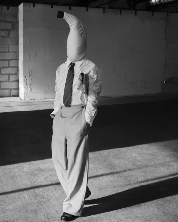 Person wearing a banana costume with a shirt, tie, and trousers stands in a concrete indoor space with a brick wall.