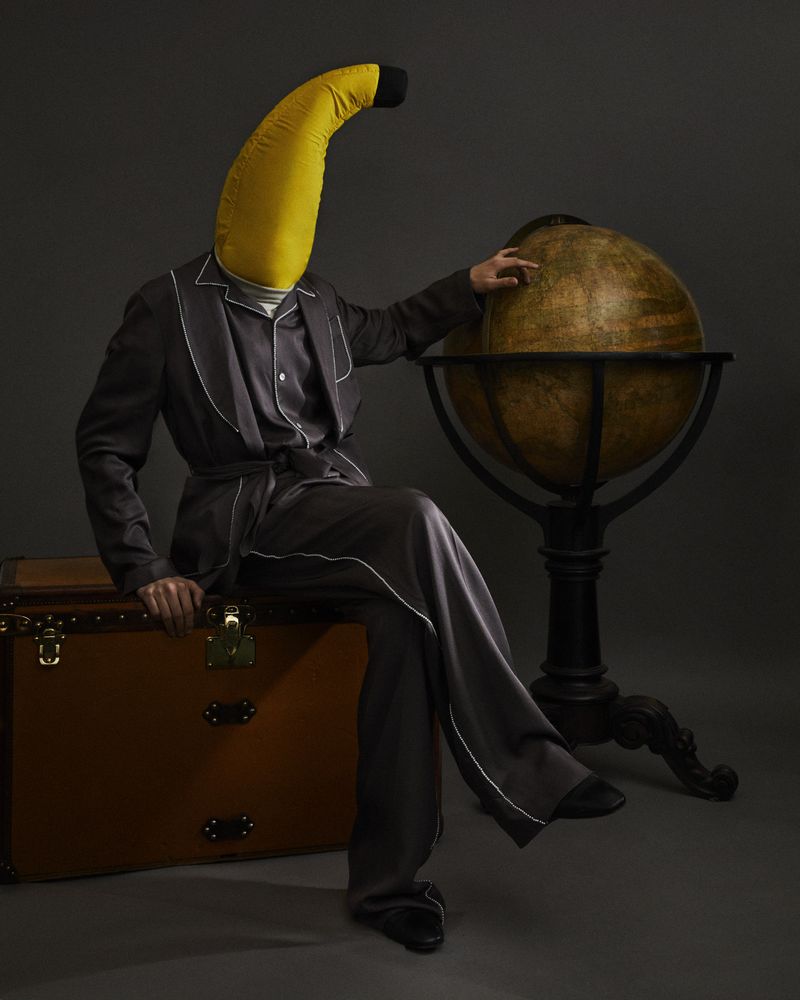 Person wearing a banana headpiece, grey pajamas with white piping, and sitting on a vintage steamer trunk next to a globe.