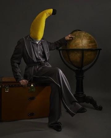 Person wearing a large banana headpiece and dark pyjamas sitting on a brown suitcase next to a vintage globe.