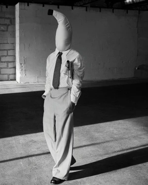 Monochrome image of a person wearing a banana mask, striped shirt, tie, and trousers in an industrial warehouse interior