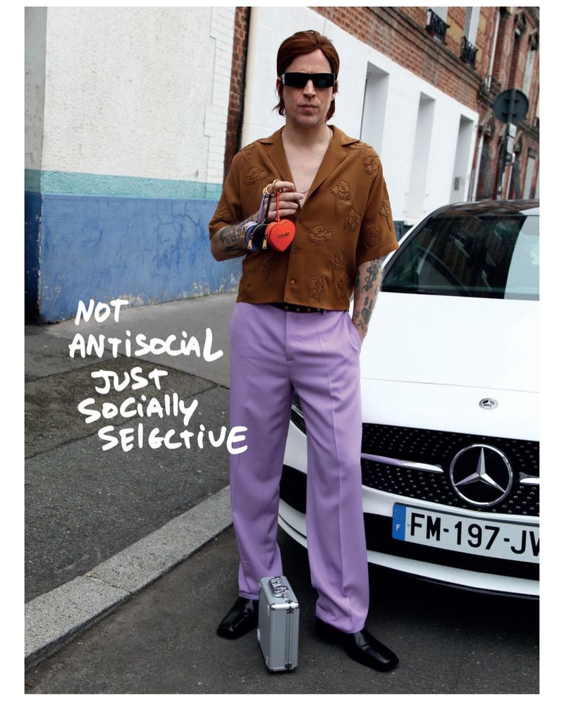 Stylish man stands in the street with a red heart-shaped purse next to a white car, wearing sunglasses, brown shirt, and lavender pants.