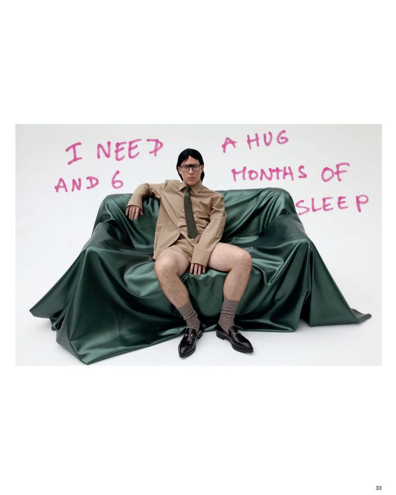 Young man in glasses sits on a draped green couch in a tan suit and tie with black shoes. Studio shot with handwritten text on the backdrop.