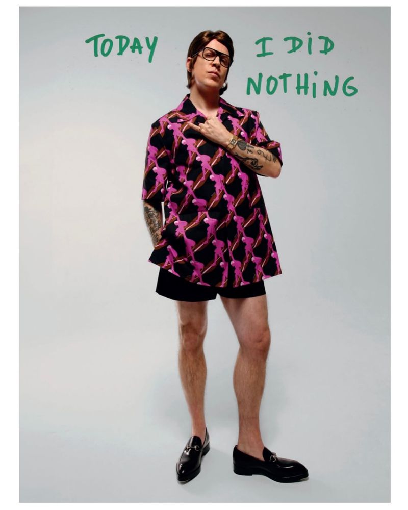 Adult male wearing a patterned shirt, shorts, and loafers stands in a studio with the text 'TODAY I DID NOTHING' on the wall.