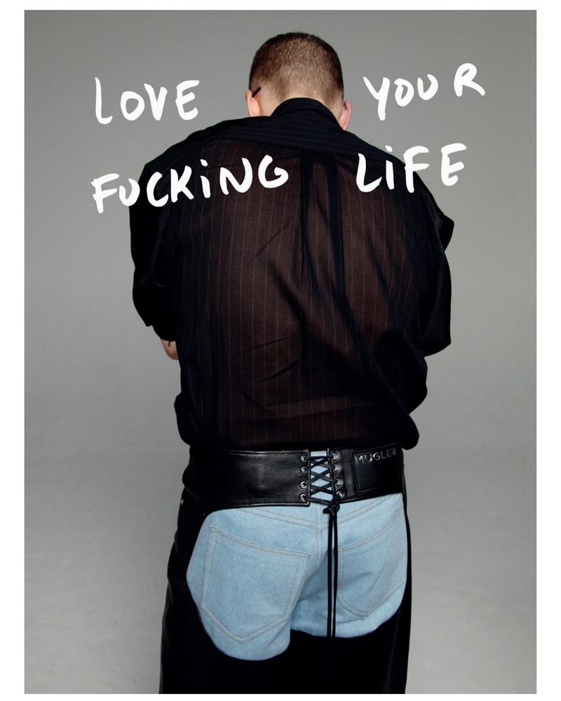 An adult male with a shaved head wears a sheer black shirt, jeans chaps, and a black belt in a studio shot with white text overlay.