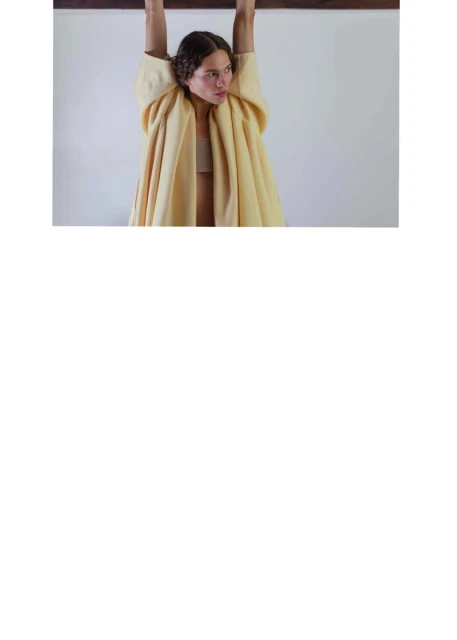 Young woman in a light yellow jacket hanging from a brown bar. Portrait shot against a white wall background. Natural light.