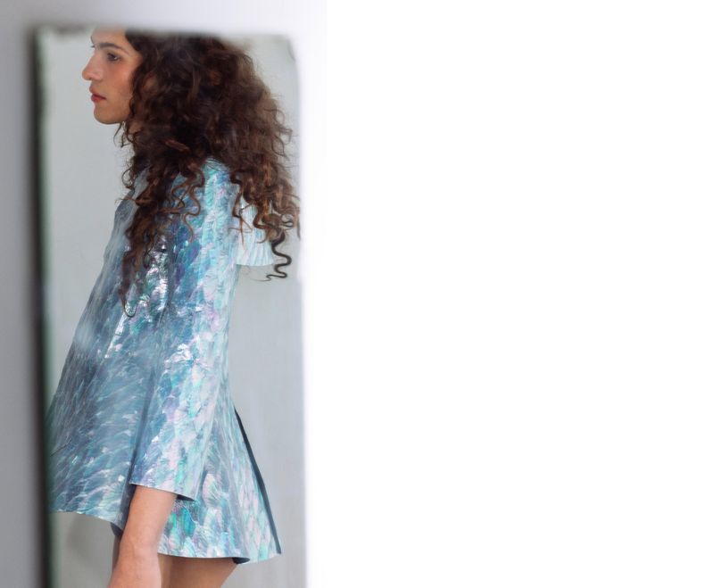 Woman in a shimmering blue mini dress with curly dark hair, captured in a light-filled indoor setting, reflected in a mirror.