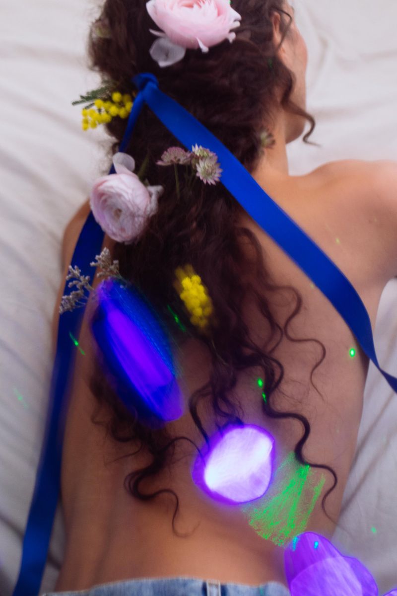 View of a woman's back with curly hair decorated with flowers and a blue ribbon against a white fabric background with bright light effects.
