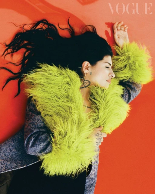Brunette woman with yellow fur collar lies on an orange background in a fashion shot. Vogue magazine logo is at the top.