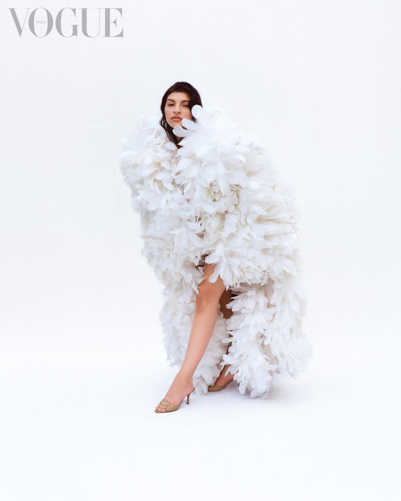 Elegant woman in a white feather gown poses in a studio. She is wearing nude heels. The Vogue logo is in the upper left corner.