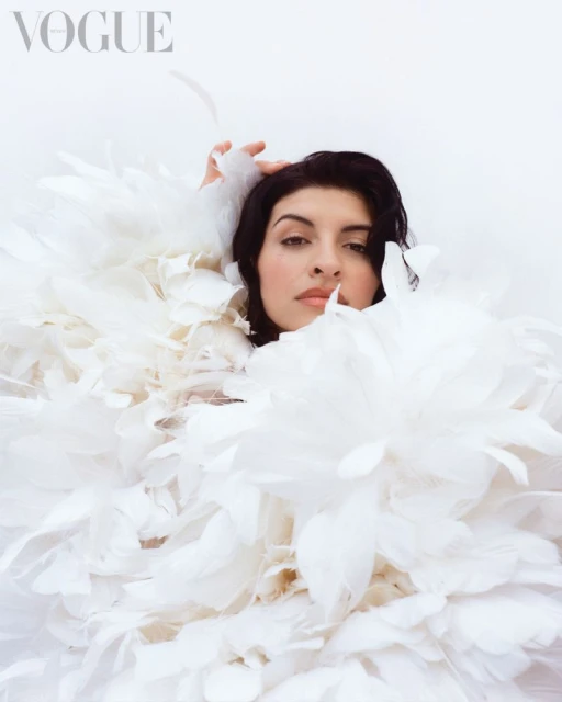 Brunette woman poses in a close-up shot wearing a white feathered dress, set against a bright white background for a Vogue fashion editorial.