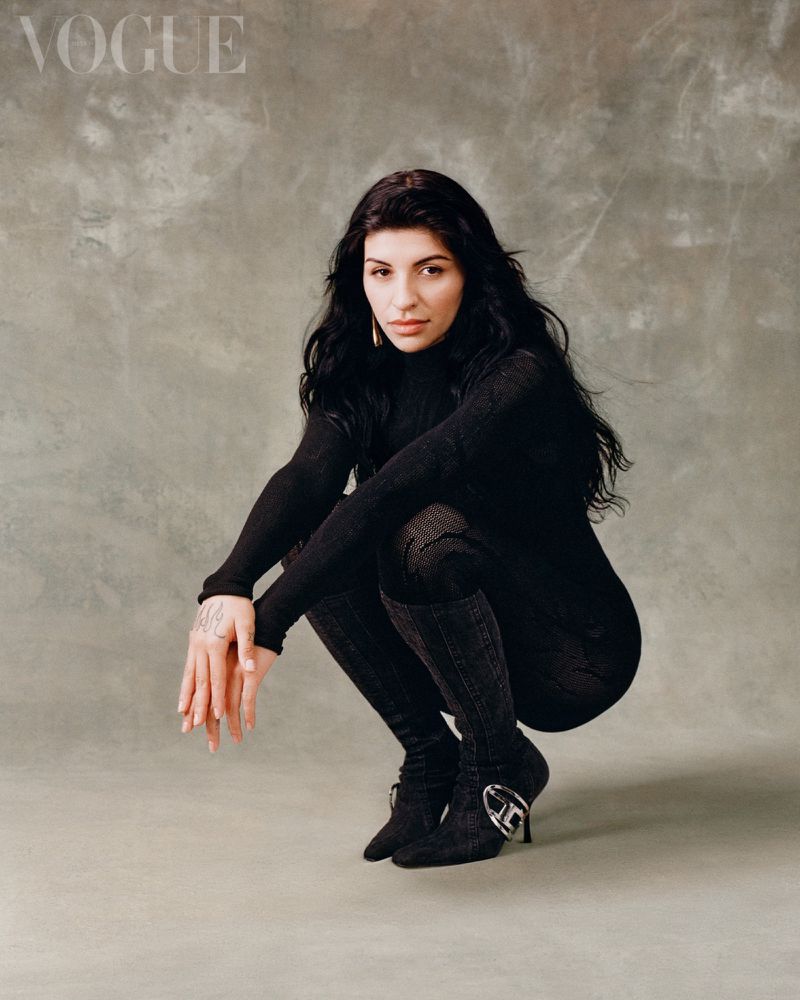 Brunette woman in black turtleneck and high boots crouches indoors, Vogue logo visible in background. Fashion editorial, portrait photography.