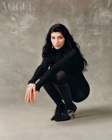 Brunette woman in black turtleneck and high boots crouches indoors, Vogue logo visible in background. Fashion editorial, portrait photography.