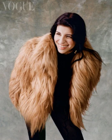 Cazzu smiling in a voluminous faux fur coat against a textured grey background for Vogue Mexico editorial.