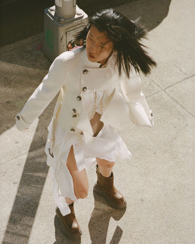 Asian woman in motion outdoors wearing white jacket, dress, and brown platform boots on sidewalk with street light shadow.