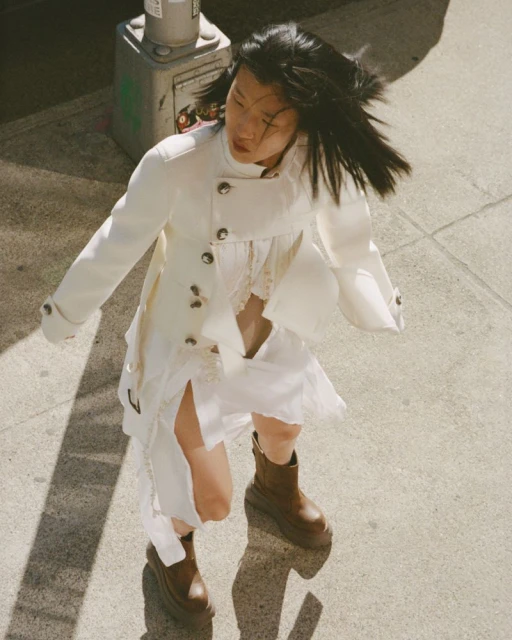 Asian woman in motion outdoors wearing white jacket, dress, and brown platform boots on sidewalk with street light shadow.