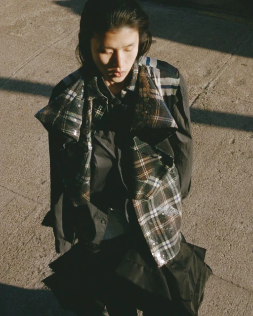 An adult woman with long dark hair in a fashionable plaid shawl stands on a sidewalk in sunlight wearing a dark-colored outfit, gazing downward.