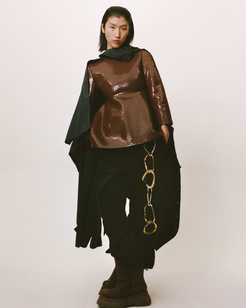 Fashion editorial shot of a stylish woman in a brown sequin top, black trousers with gold chain, and boots against a neutral backdrop.