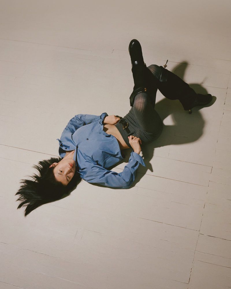 A young adult woman with long black hair lays on a white surface, wearing a blue shirt, grey patterned shorts, and black boots.