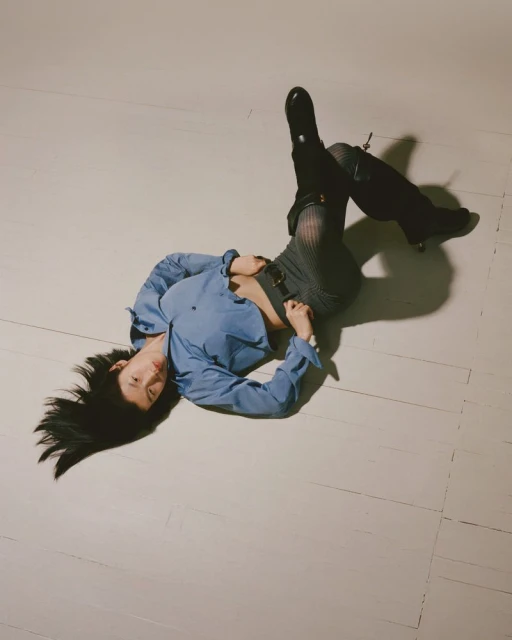 A young adult woman with long black hair lays on a white surface, wearing a blue shirt, grey patterned shorts, and black boots.