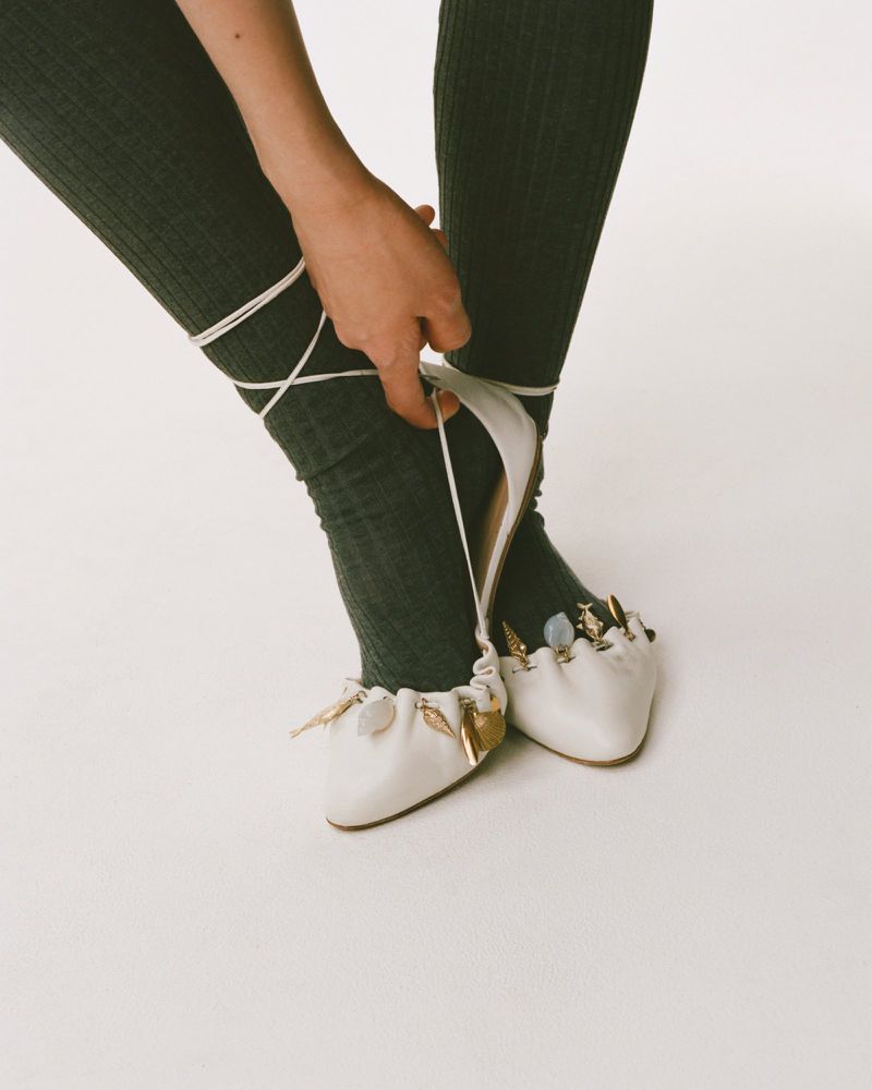 A person fastens the straps of a pair of cream-colored ballet flats, accessorized with golden leaves, indoors against a plain background.