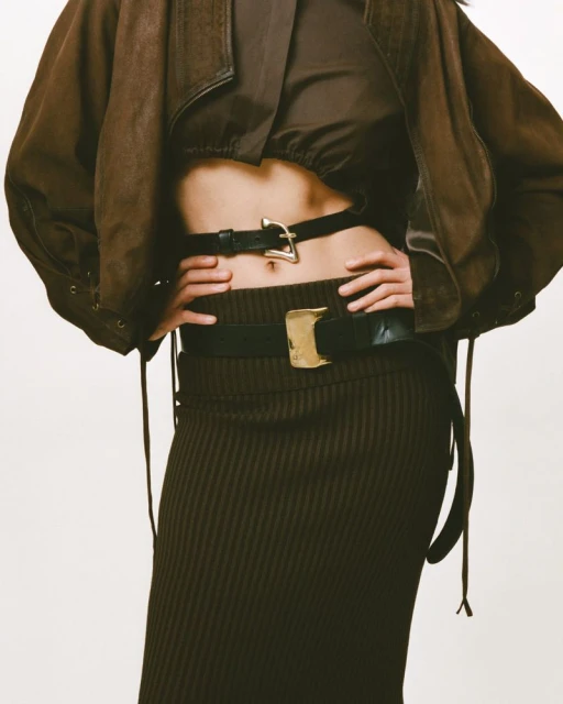 Fashionable woman posing in brown crop top, jacket, and skirt with black belts against a white background in a studio setting.