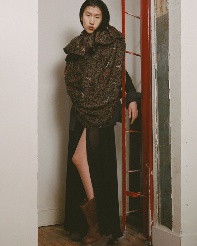 Asian female fashion model in black skirt and boots, stands near a red ladder wearing a floral jacket in a studio environment.