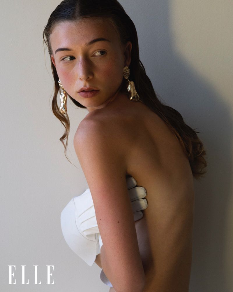 Young adult woman poses partially nude in front of a light wall, adorned with gold earrings. Her arm supports a white textile piece.