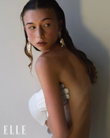 Young adult woman poses partially nude in front of a light wall, adorned with gold earrings. Her arm supports a white textile piece.