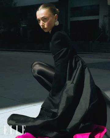 Fashion shot of adult woman crouching in black dress with pink lining, black tights, and statement earrings; dark urban setting with bright sunlight.