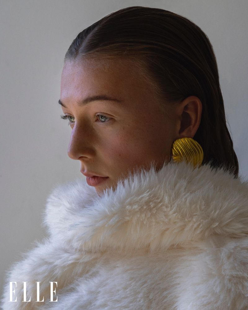 Close-up portrait of a model with wet slicked-back hair, wearing gold earrings and a fuzzy white coat, posing indoors with natural lighting.