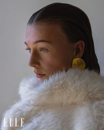 Close-up portrait of a model with wet slicked-back hair, wearing gold earrings and a fuzzy white coat, posing indoors with natural lighting.